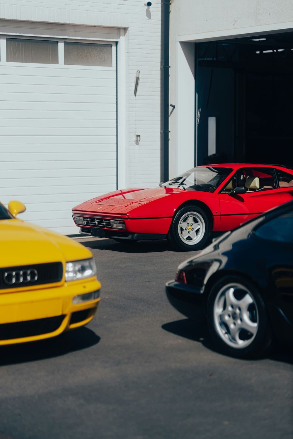 A red classic sports car parked outside a garage, a yellow car beside it